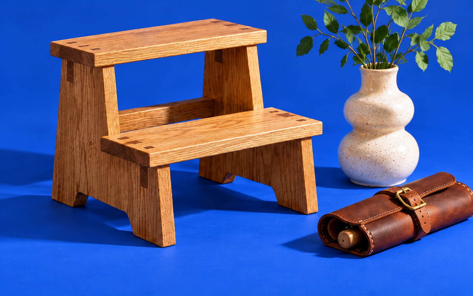 Handcrafted oak step stool, a ceramic vase, and a leather tool roll against a blue backdrop